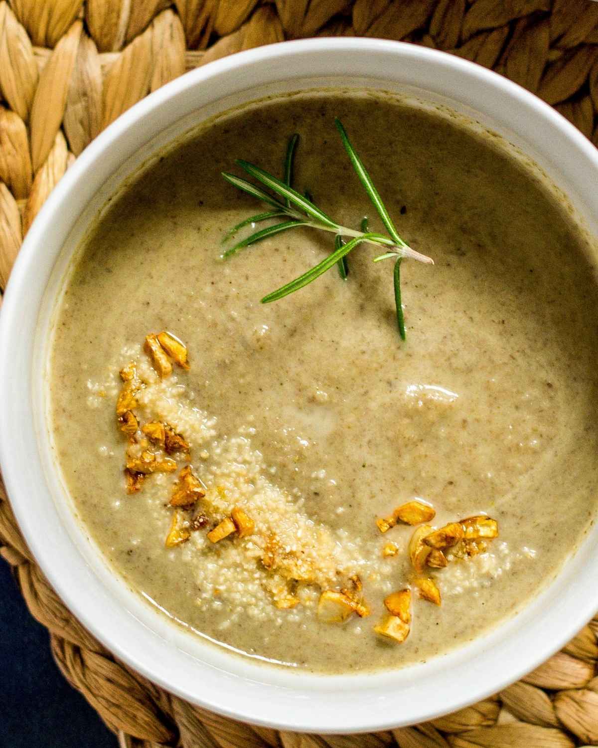 a bowl of soup with garnish of rosemary and garlic on a brown rattan mat 