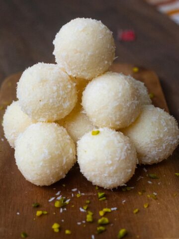 A stack of coconut ladoos piled on a wooden board with scattered pistachios