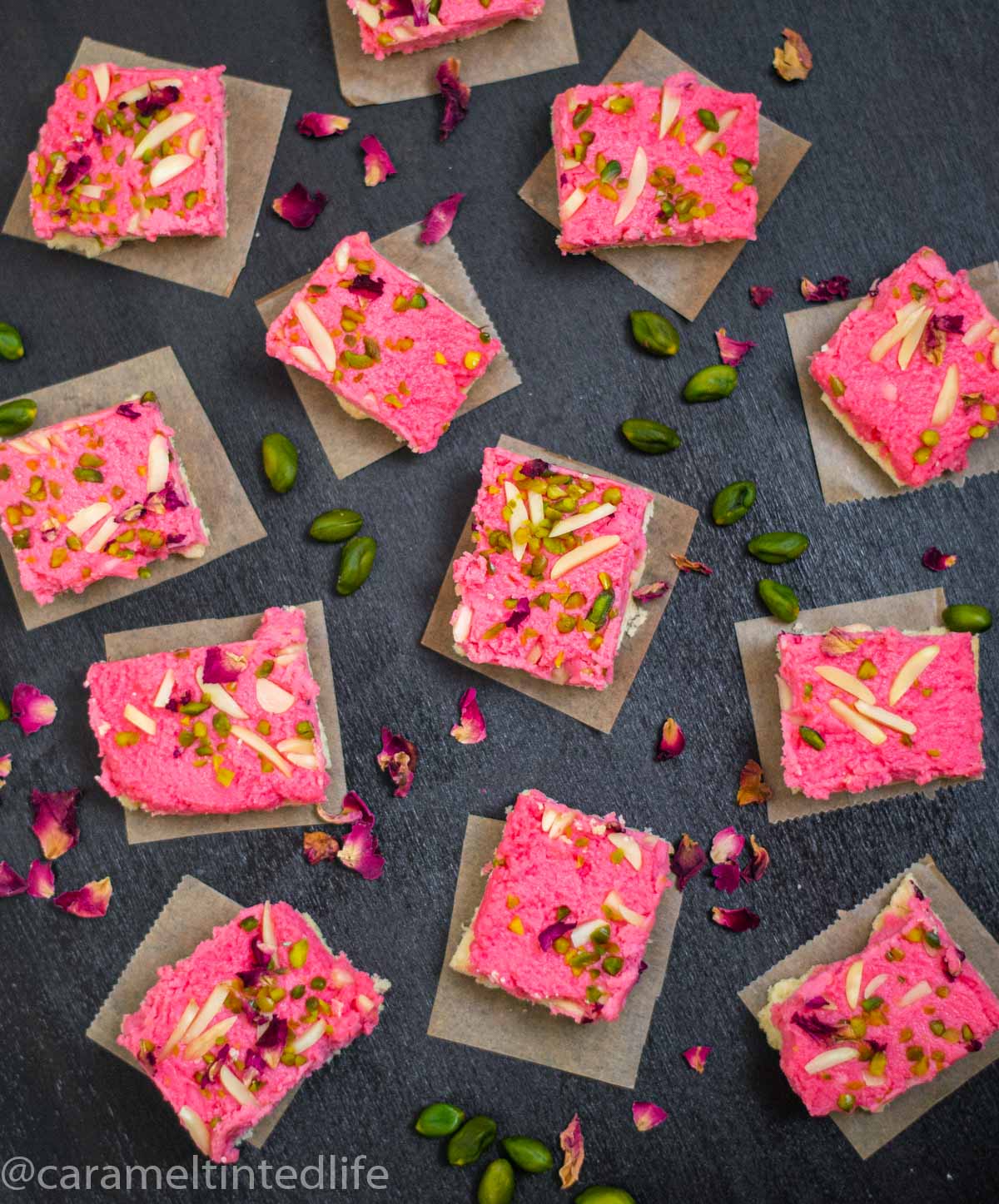 Kalakand squares on a black background viewed from the top, with scattered pistachios and rose petals