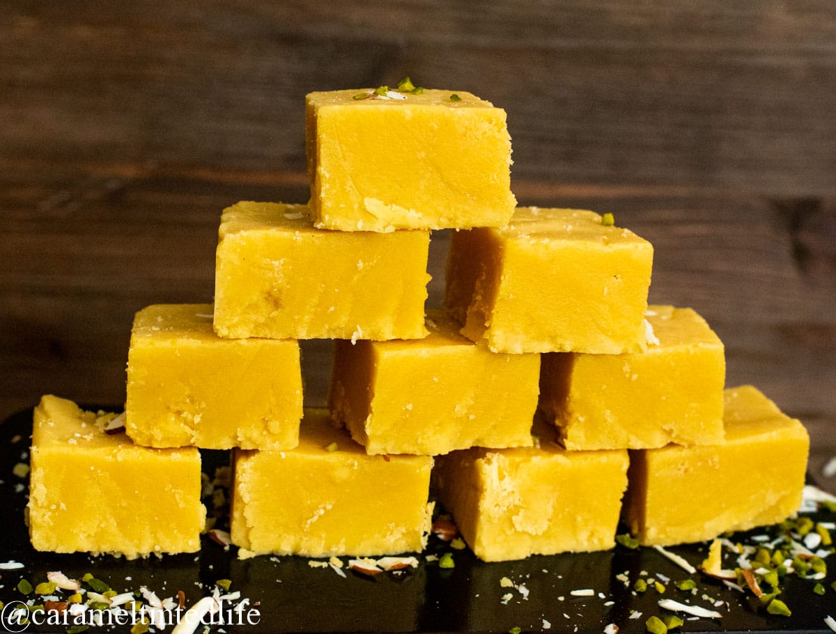 Mysore pak pices stack like a bricks on a black baking tin, against a wooden backdrop