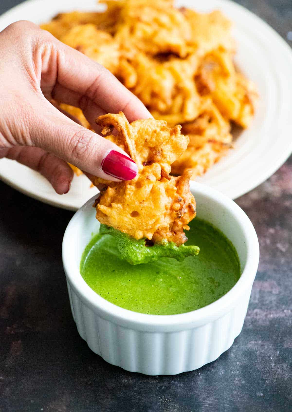 Fingers holding a fritter dipped in a white ramekin filled with green chutney