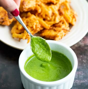 Hand holding a spoon dipping in a white ramekin filled with green chutney