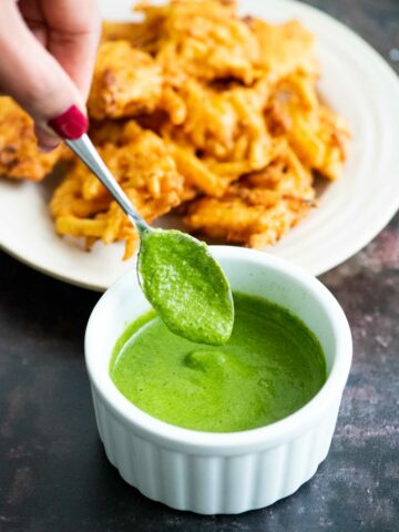 Hand holding a spoon dipping in a white ramekin filled with green chutney