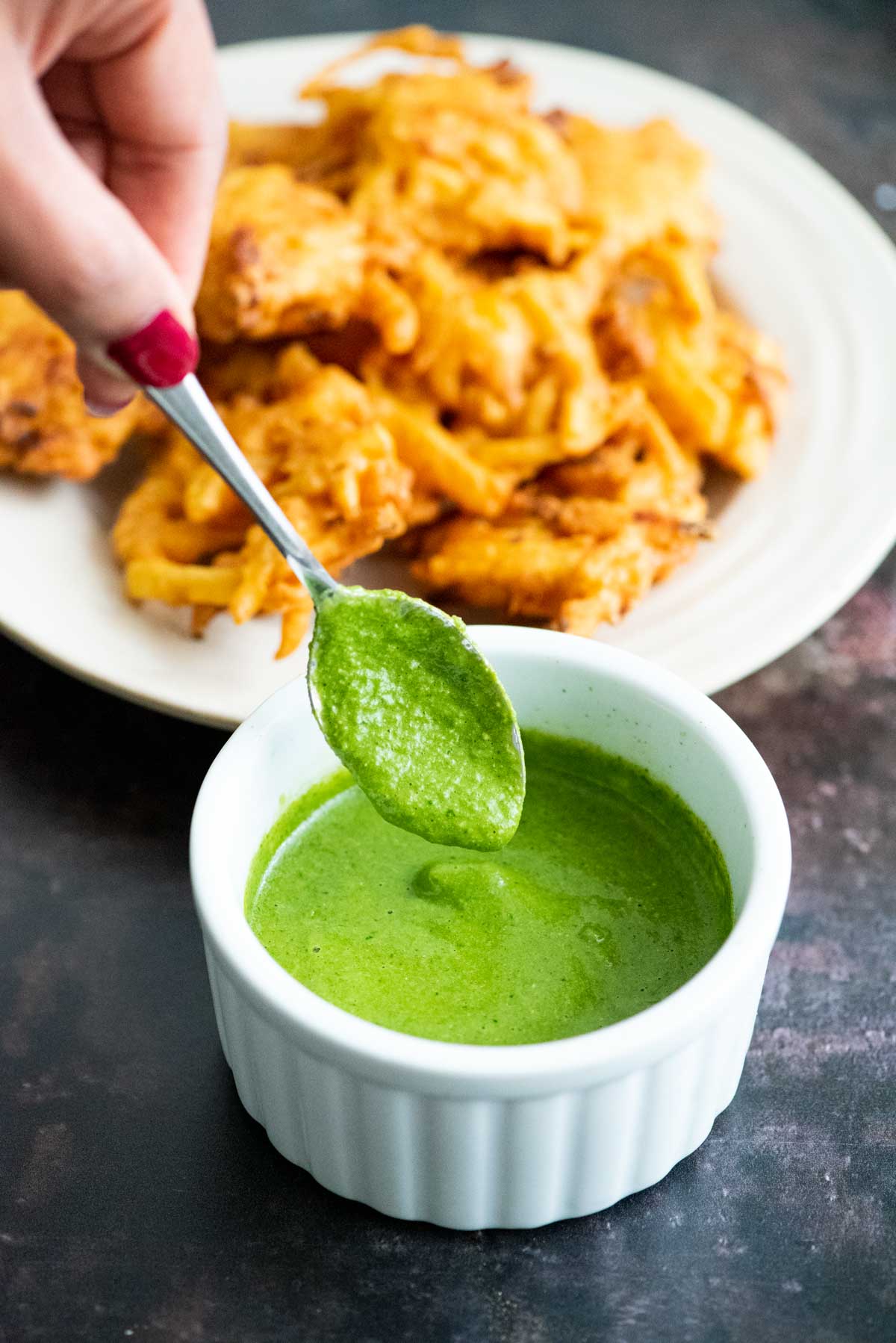 Hand holding a spoon dipping in a white ramekin filled with green chutney