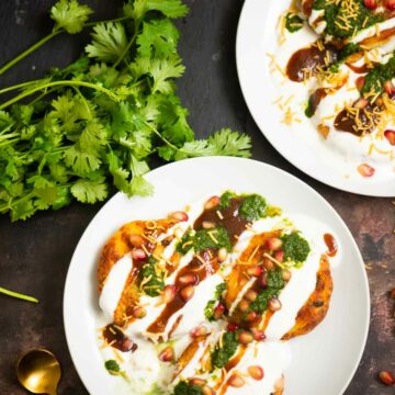 two plates of aloo tikki chaat, cilantro, and a spoon on a brown backdrop