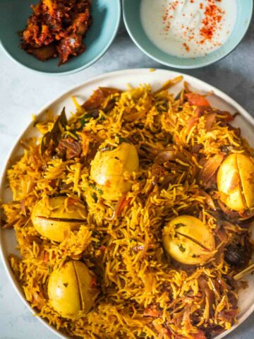 egg biryani on a plate with pickle and yogurt on the side
